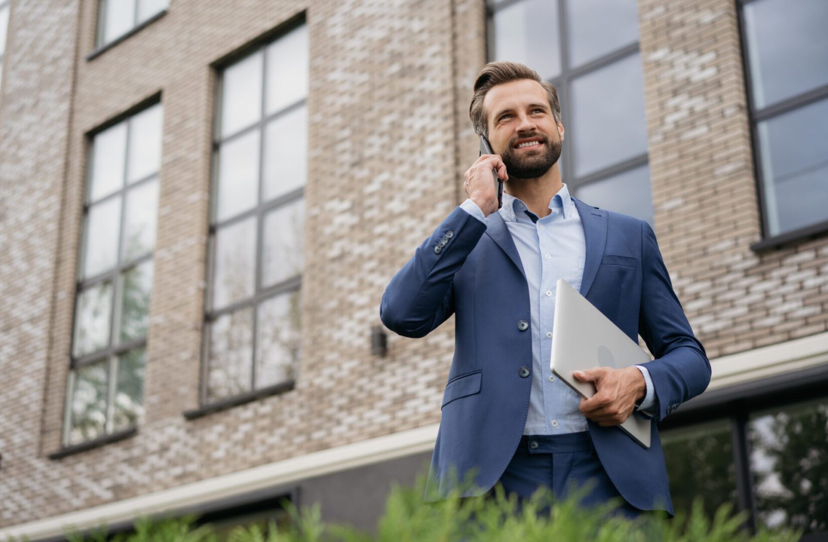 Businessman wearing stylish suit talking on mobile phone, holding laptop standing on the street. Successful business Man in suit talking on phone outside.
