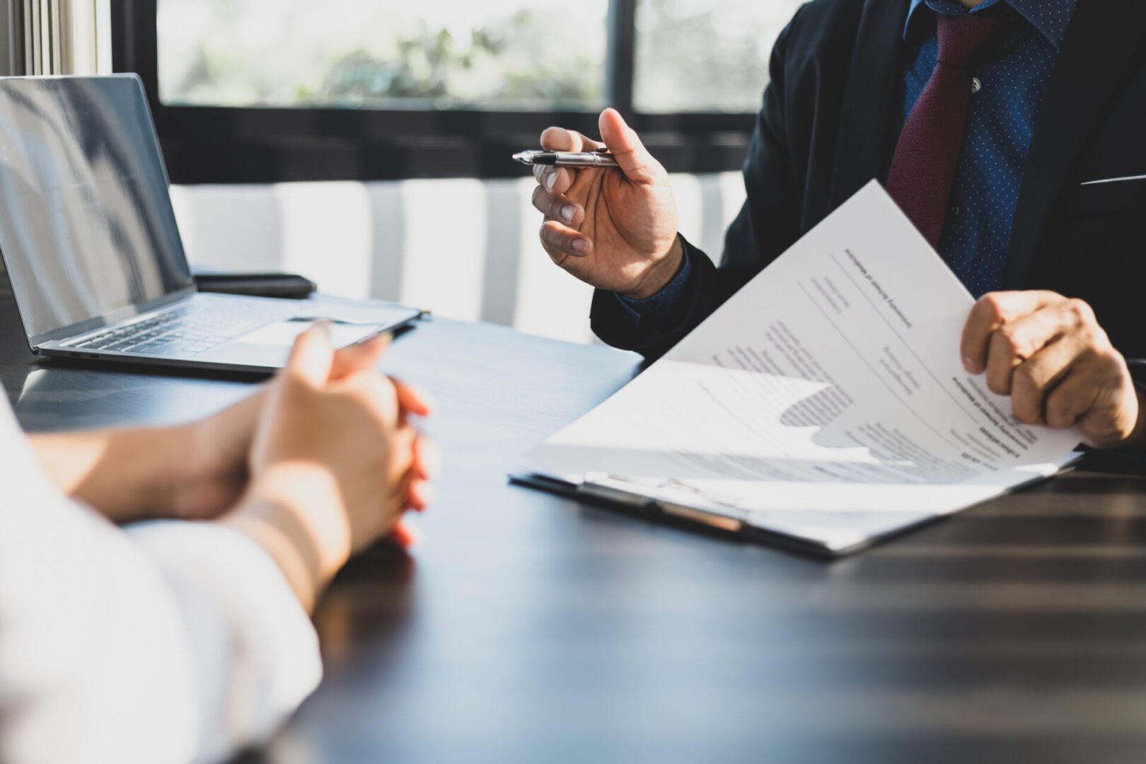 Businessman in suit in his office showing an insurance policy and pointing with a pen where the policyholder must to sign. Insurance agent presentation and consulting insurance detail to customer. Business meeting with documents and laptop.
